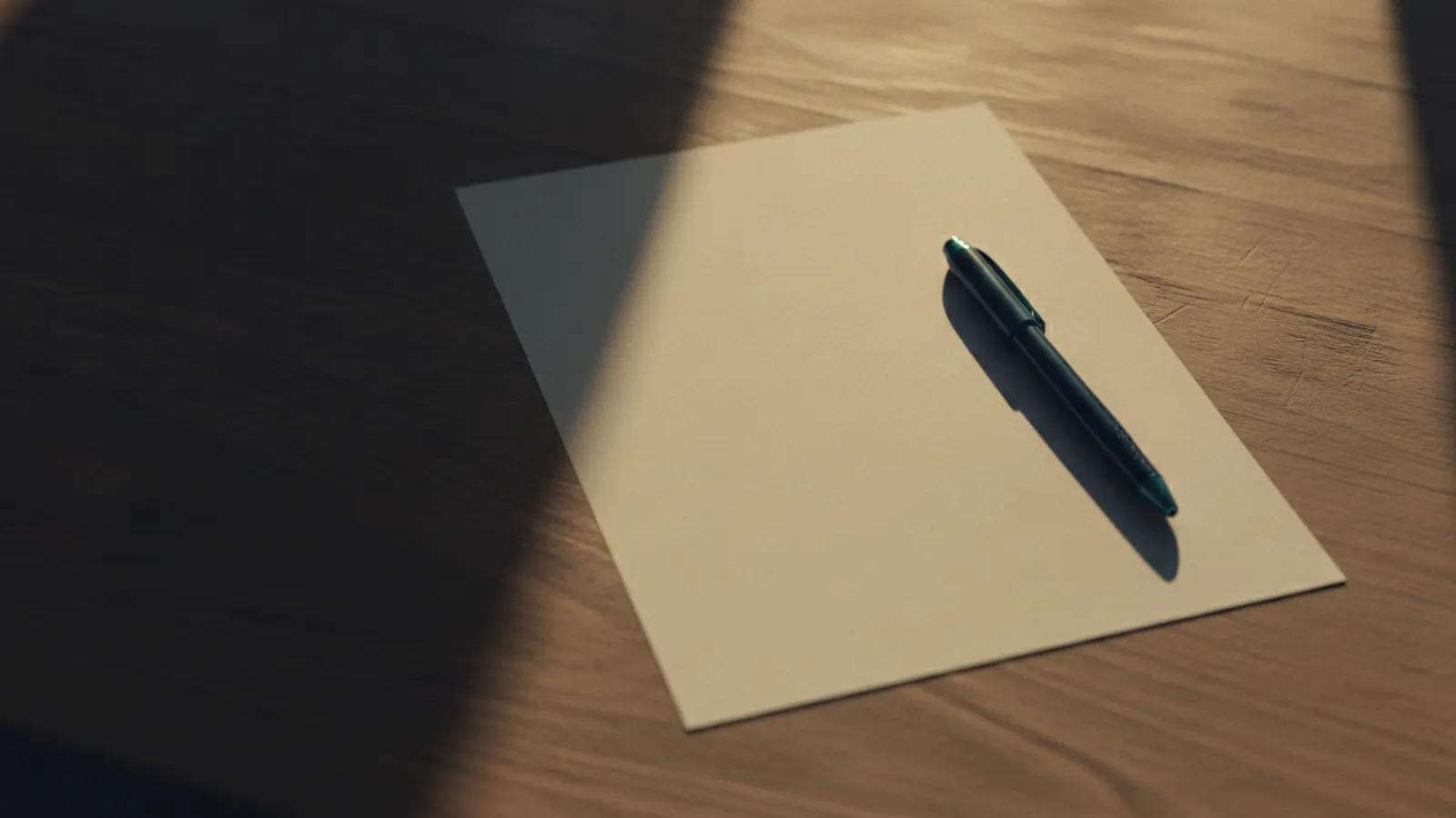 A single blank sheet of paper on a bare wooden table in low golden light, an uncapped pen beside it casting a long shadow.